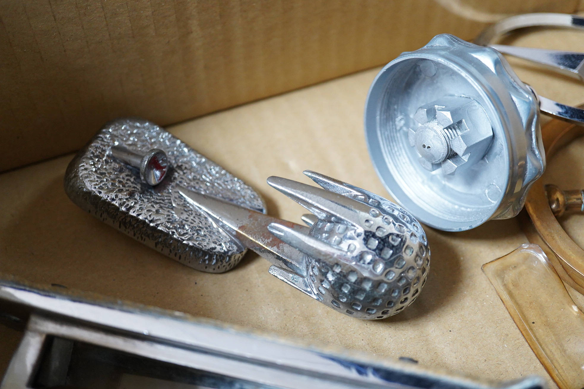 Seven chrome and brass car mascots, including a Mercedes-Benz logo, a figure of a blacksmith, a caricature figure on a beer barrel, a golf ball in mid flight, etc. together with a chromed housing unit for an electric lam